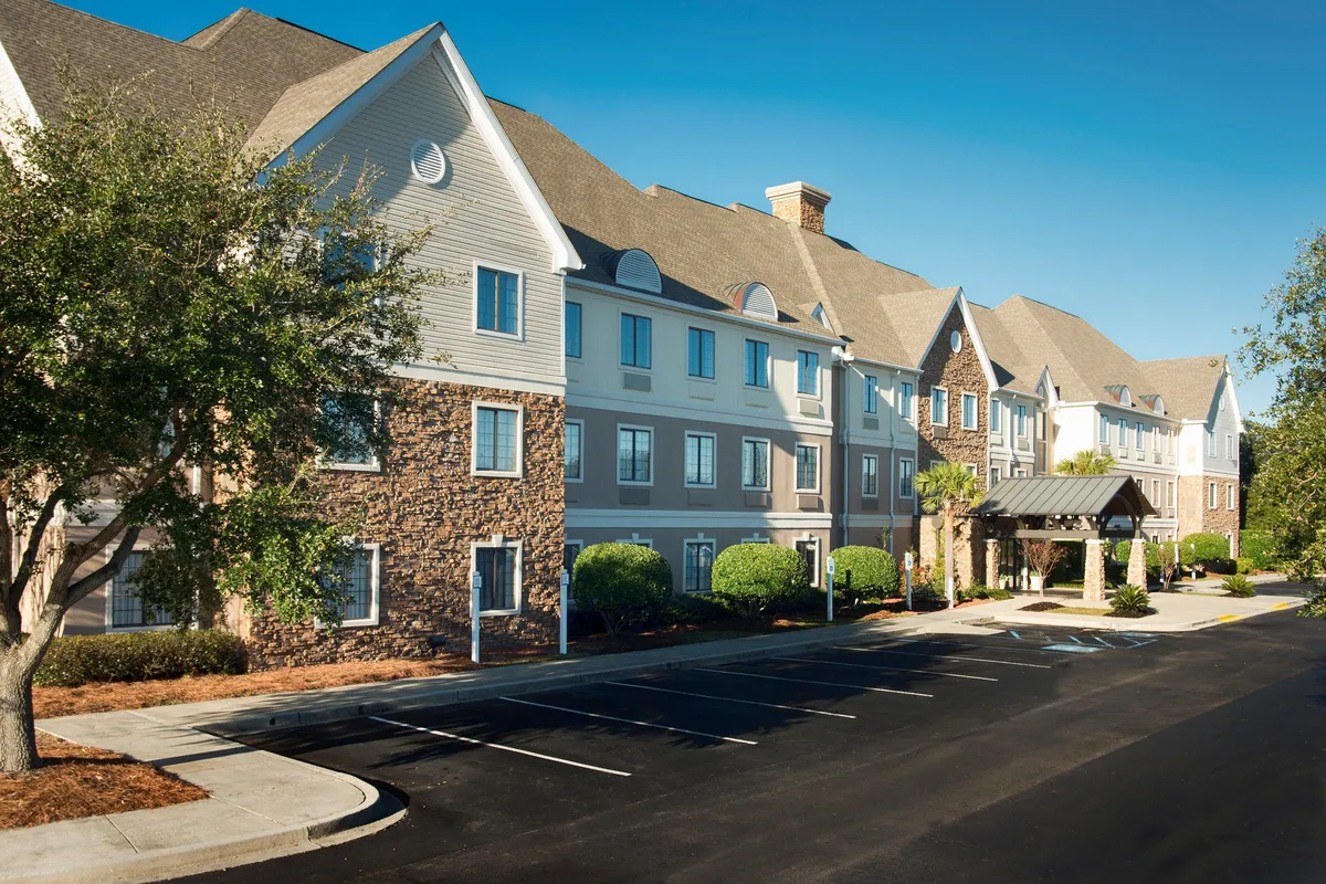 This image features the inviting entrance canopy of the hotel, set against a stone facade and complemented by well-maintained landscaped shrubs. The setting is comfortable and approachable, framing the arrival experience and hinting at ample on-site parking.