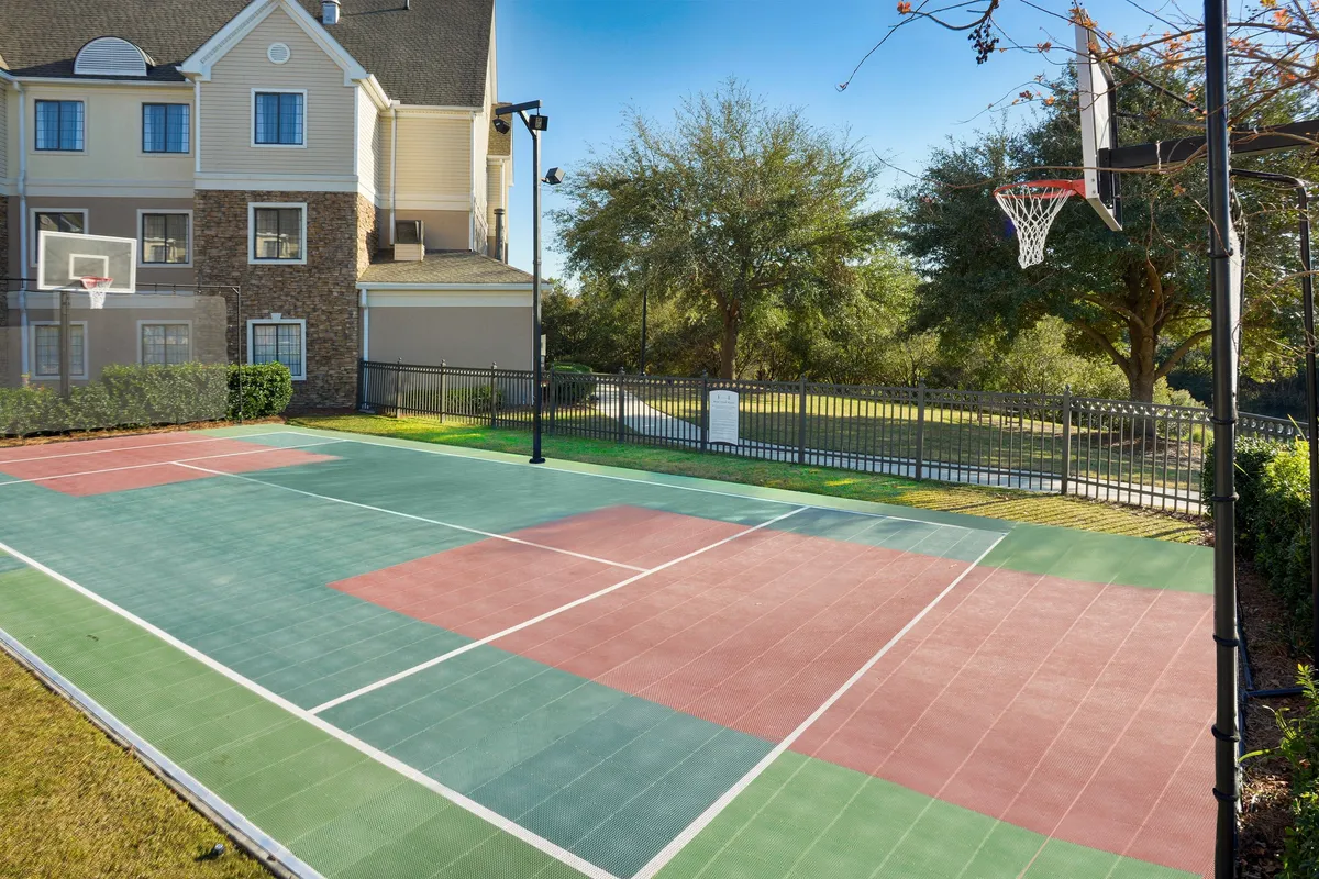 A vibrant outdoor basketball court surrounded by lush greenery, with a prominent basketball hoop in the foreground. The scene suggests friendly matches and active recreation in a pleasant setting.