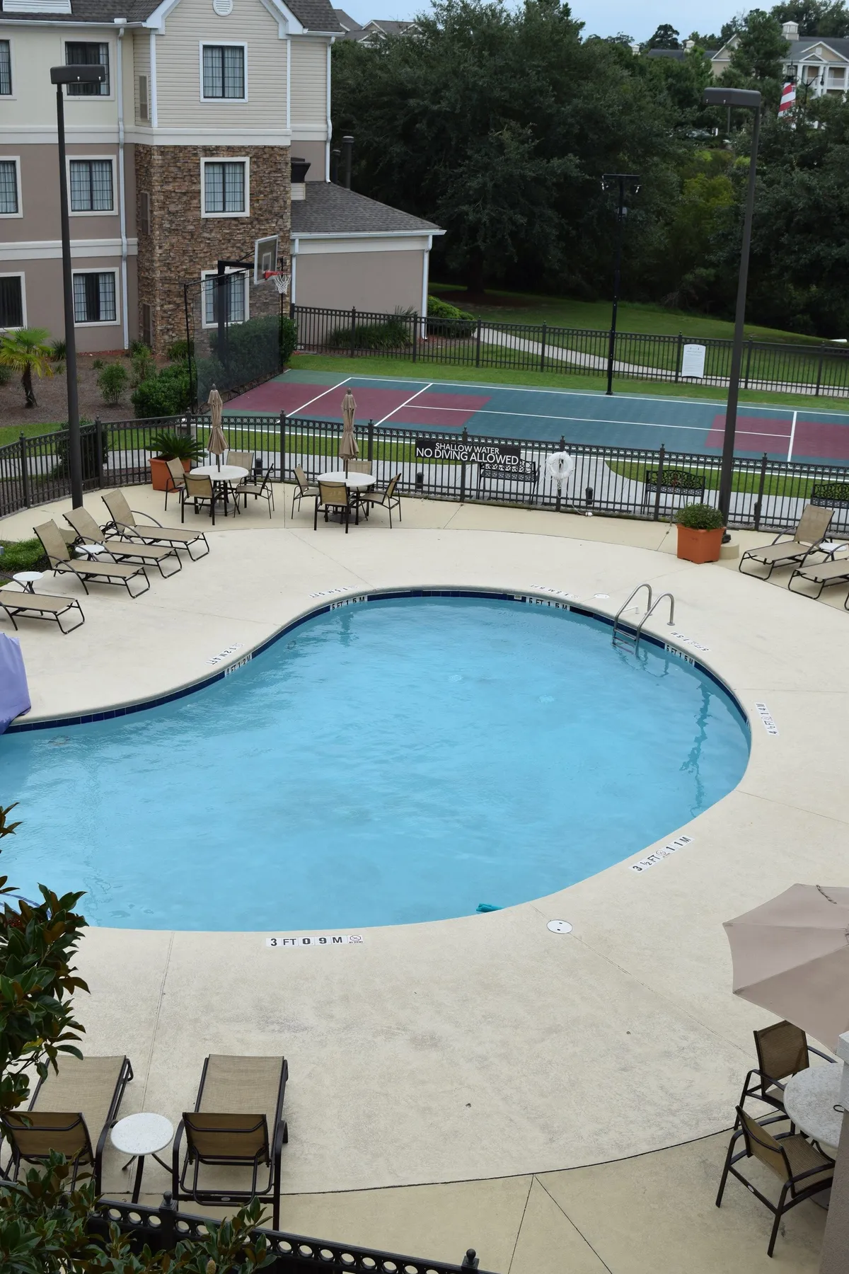The scene showcases a vibrant outdoor area featuring a large swimming pool surrounded by lounge chairs and a tennis court, with the resort building in view. This lively setting encourages active family time, whether swimming, sunbathing, or playing sports together.