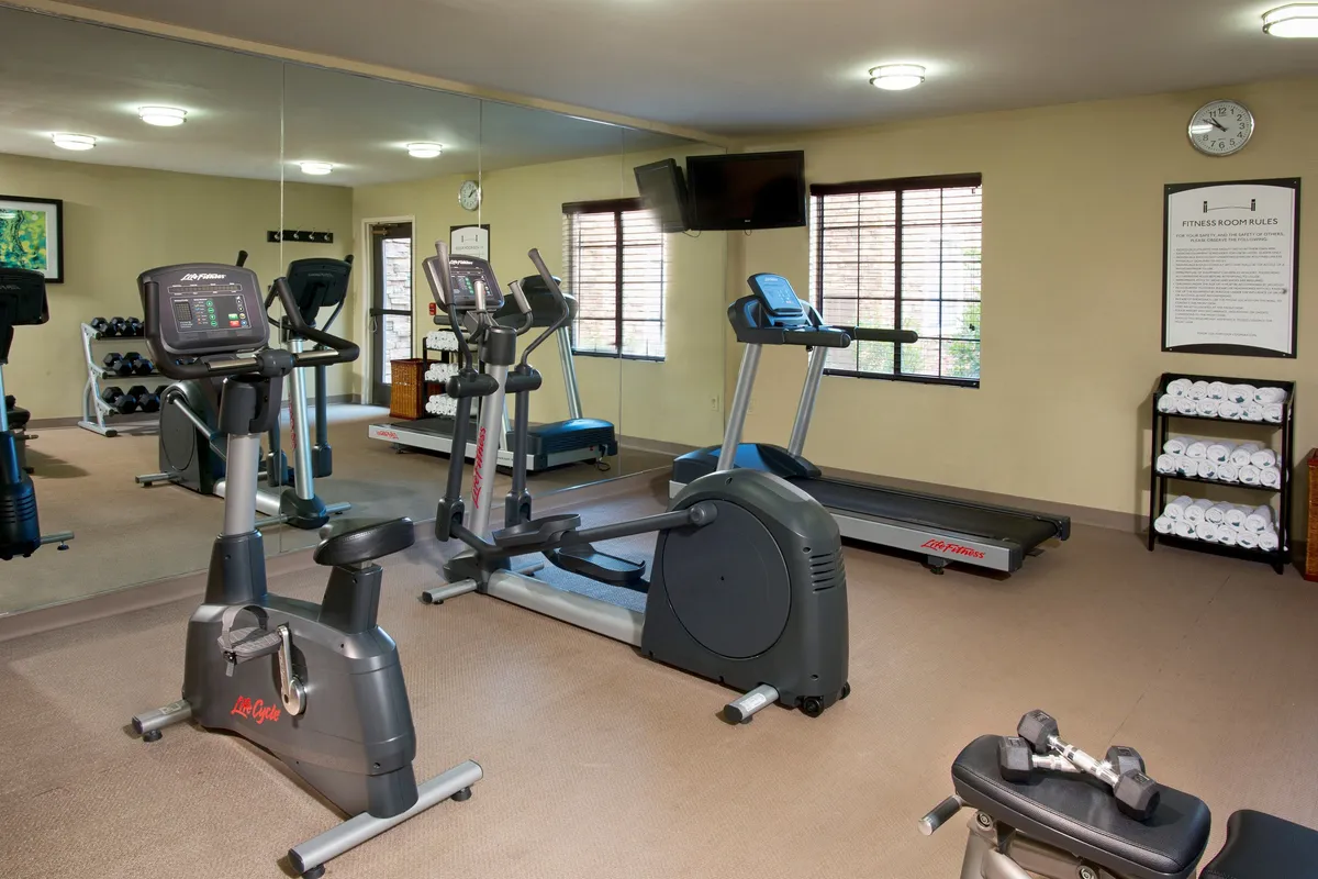An energetic, modern fitness center equipped with a stationary bike, treadmill, weights rack, and a mirrored wall. The space is well-lit, ideal for both solo fitness routines and group workouts.