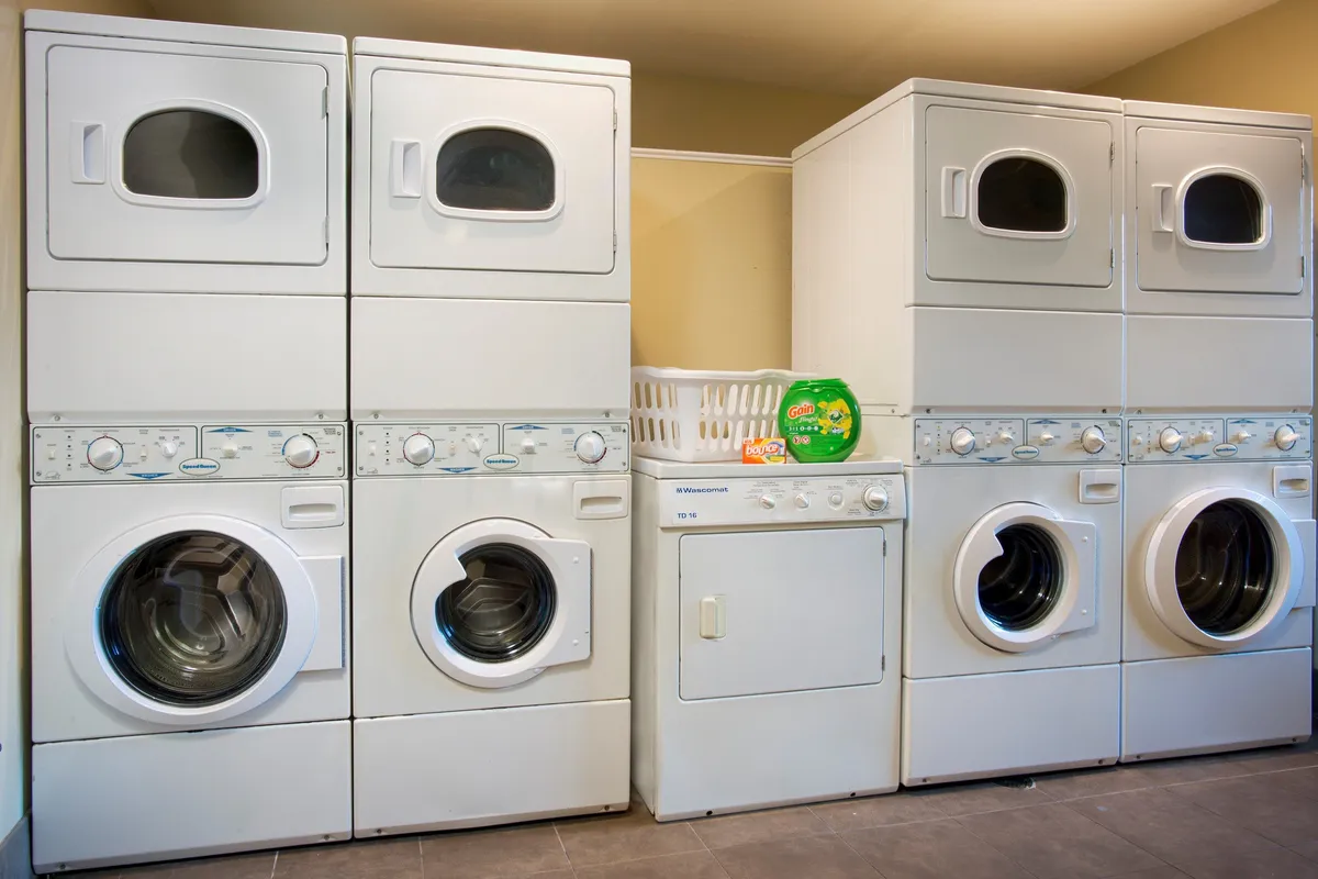 A compact laundry area featuring stacked washer and dryer units, a laundry basket, and detergent. The setting is practical and uncluttered, directly illustrating the availability of on-site laundry facilities for personal convenience.
