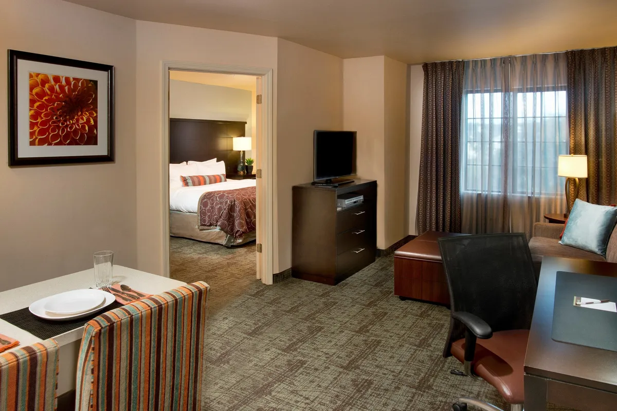 The hero image showcases a cozy and modern condo interior, featuring a welcoming dining area set for a meal, a glimpse into the inviting bedroom, a sleek television unit for entertainment, and large windows with curtains that bathe the space in natural light. The layout hints at spaces perfect for dining, relaxing, working remotely, or unwinding after a day at the beach.