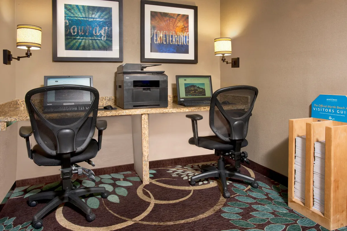 This image showcases two computer desks equipped with office chairs and a printer, complemented by motivational artwork on the wall. The setup is clearly tailored for business use, providing a practical workspace for guests needing to print documents, work efficiently, or plan activities.