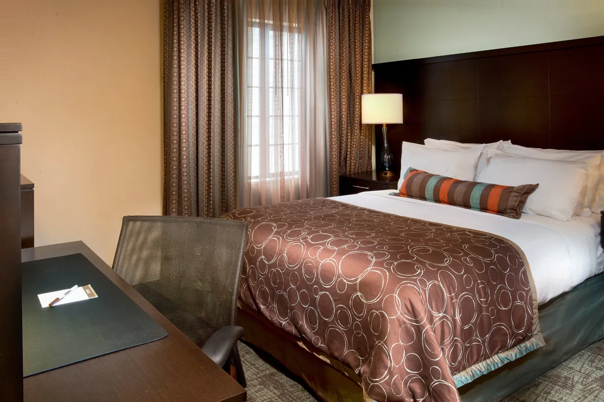 A bright and cozy bedroom featuring a queen bed with patterned bedding, a desk with writing materials, and a large window streaming in natural light. The room exudes calm and comfort, perfect for relaxation or focused work.