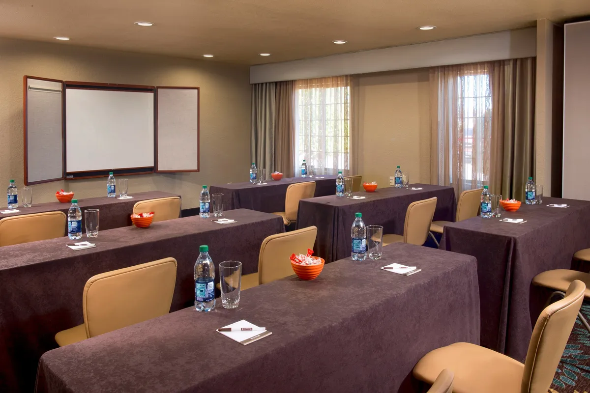 A bright, functional meeting space with long tables arranged in rows, water bottles, snack bowls, and a presentation board at the front. The room is set up for group meetings, workshops, or brainstorming sessions, reflecting a collaborative and focused atmosphere.